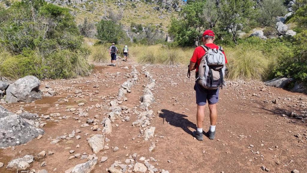 CLOT DE DÉU Y COLL DE MOIXARRINS - Caminando por Mallorca Grupos de senderistas avanzando por un camino de tierra con piedras y vegetación en el fondo, en una zona montañosa de Mallorca.