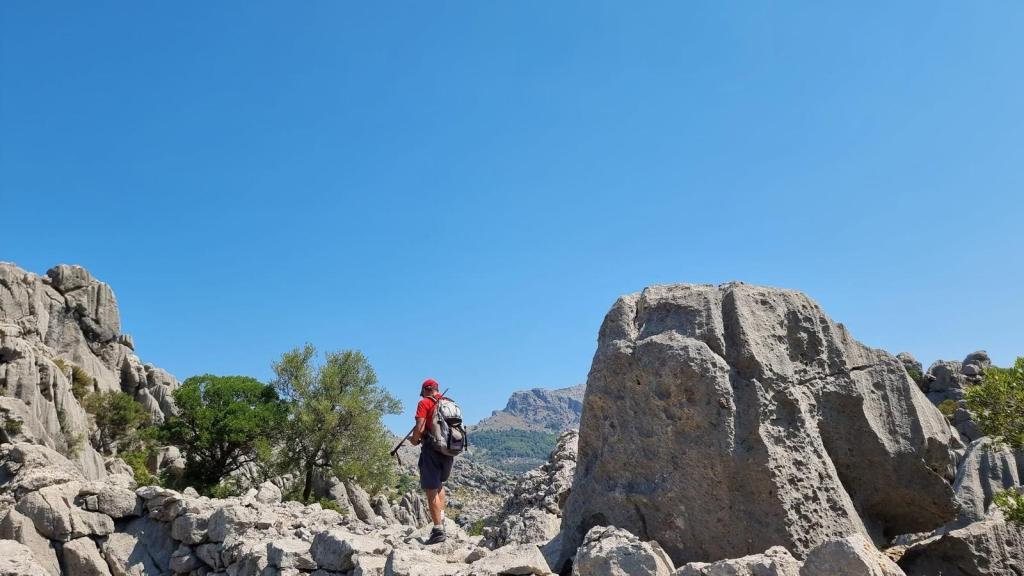 CLOT DE DÉU Y COLL DE MOIXARRINS - Caminando por Mallorca Hombre caminando entre rocas en un paisaje montañoso bajo un cielo azul despejado.
