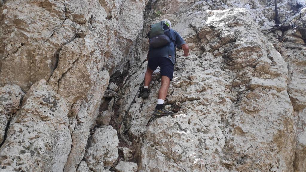 Una persona escalando una pared de roca en un sendero montañoso, con un paisaje rocoso al fondo.