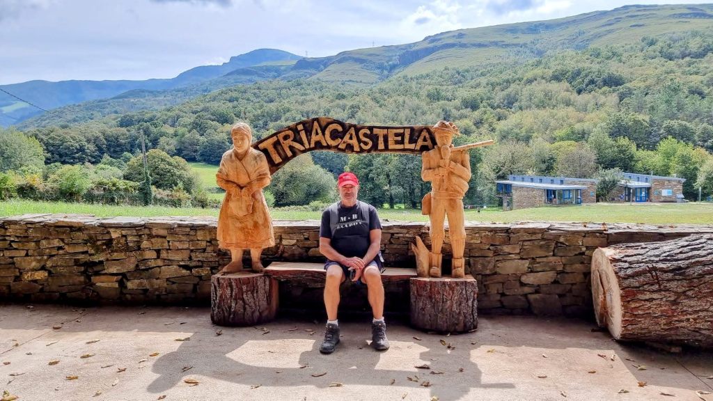 Un hombre sentado en un banco de madera con esculturas de madera representando a una mujer y un hombre detrás de él, en un entorno natural con montañas al fondo y el cartel que dice 'Triacastela'.