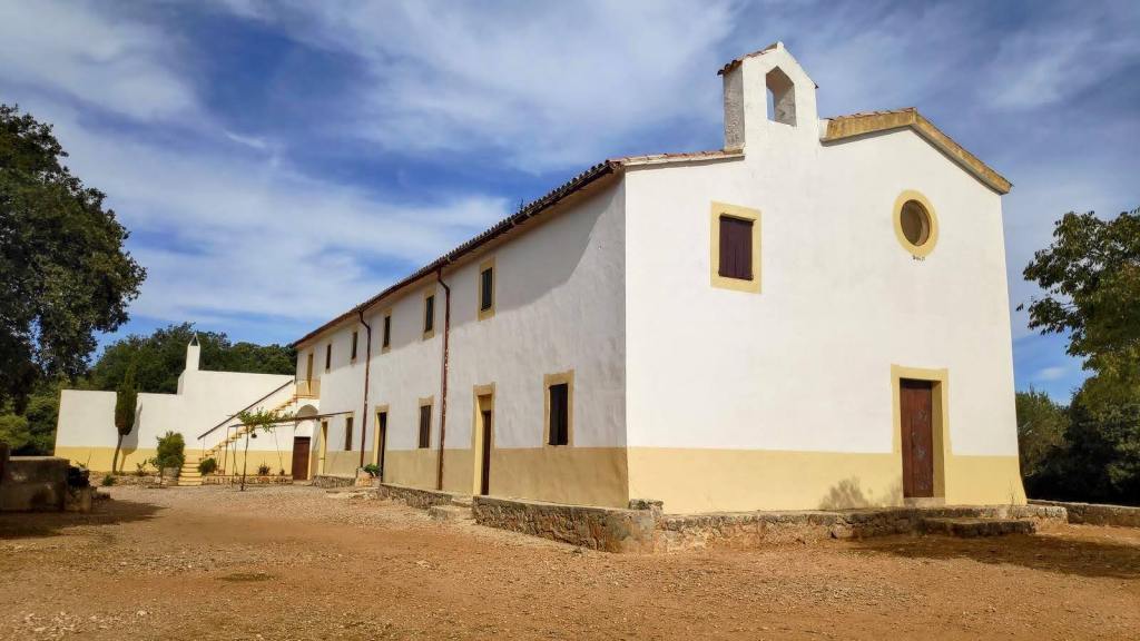Vista exterior de una edificacion blanca con detalles en color amarillo, ubicada en un entorno natural, con árboles alrededor y un cielo parcialmente nublado.