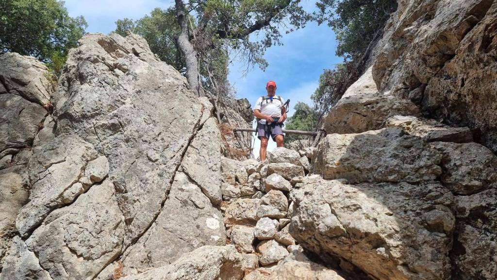 Hombre caminando por un sendero rocoso, rodeado de grandes piedras y vegetación, en una ruta de senderismo.