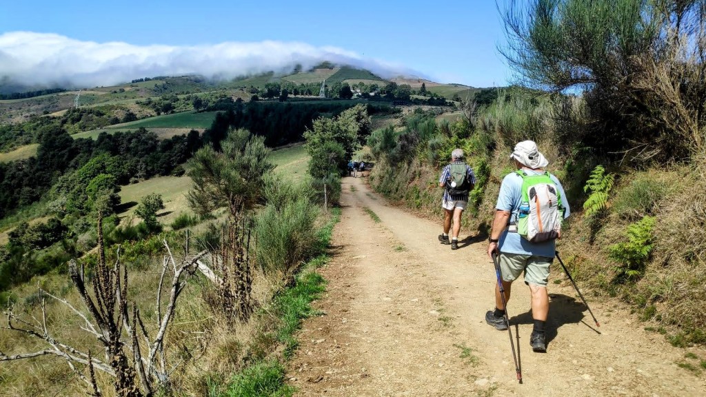 Dos peregrinos caminando por un sendero rural rodeado de vegetación y montañas, con nubes bajas en el horizonte.