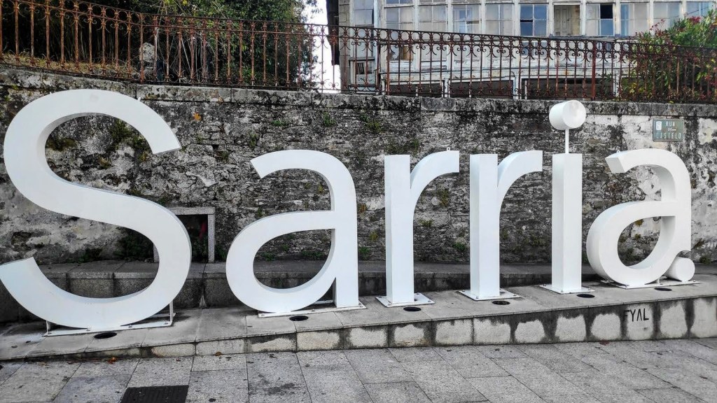 Cartel blanco con la palabra 'Sarria' en letras grandes, ubicado en un espacio público rodeado de una pared de piedra.