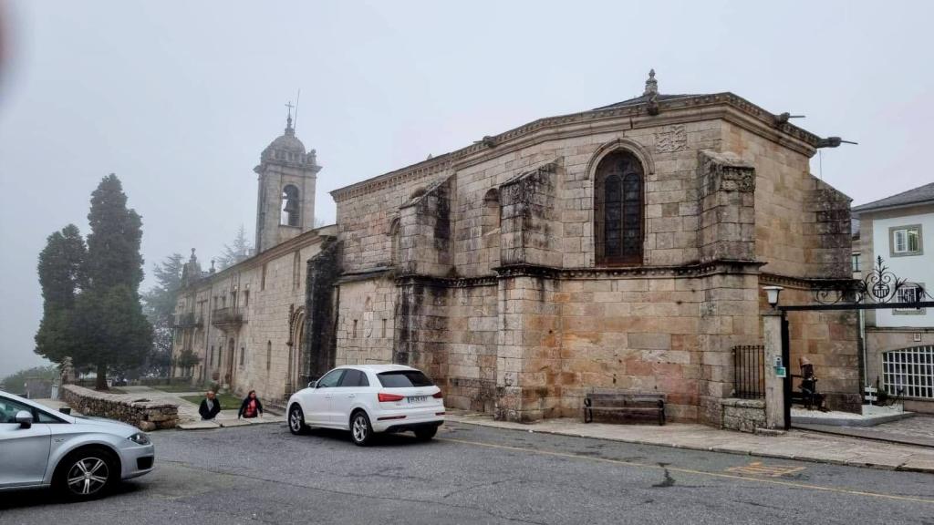 Vista de un edificio histórico con una torre campanario, rodeado de árboles y autos, en un ambiente nublado.