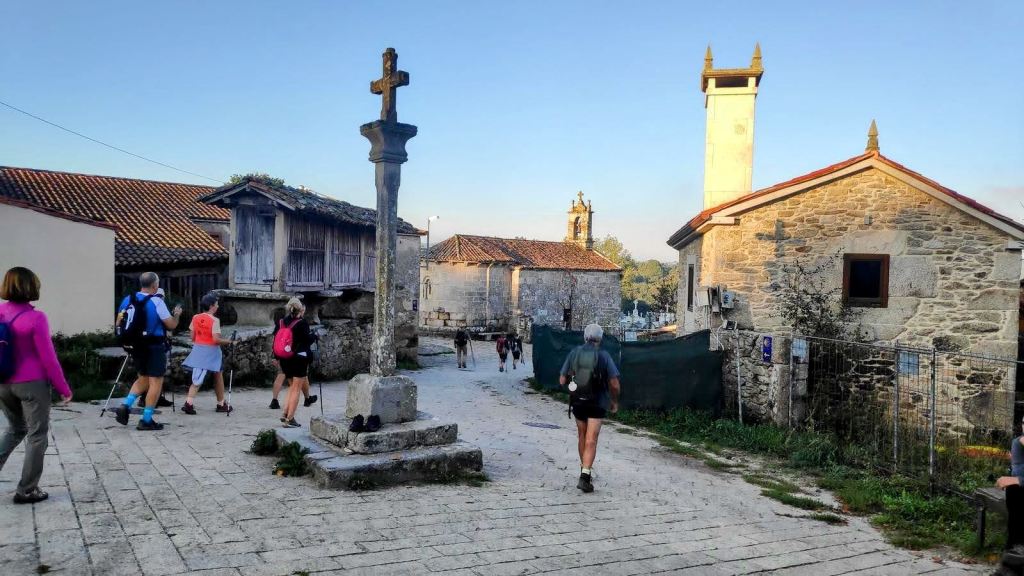 Grupo de peregrinos caminando por una calle empedrada en una aldea del Camino de Santiago, con una cruz en el centro y hórreos de fondo.