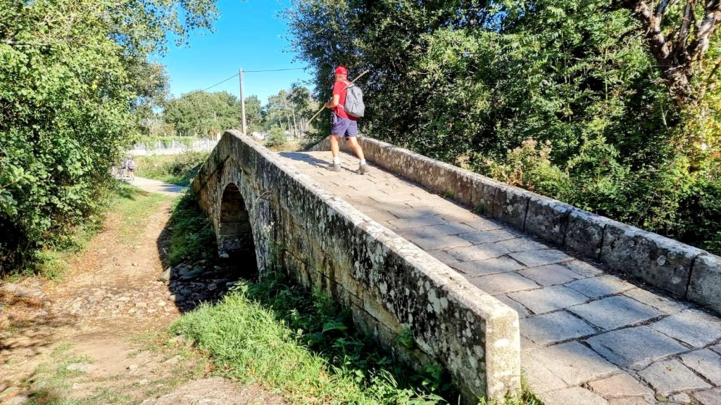 Peregrino cruzando un puente de piedra en el Camino de Santiago, rodeado de vegetación y un cielo despejado.