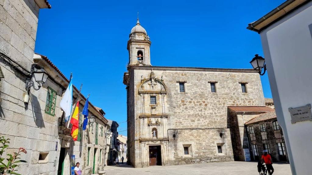 Vista de una iglesia de piedra con una torre en un día soleado, rodeada de edificios tradicionales en una calle tranquila.