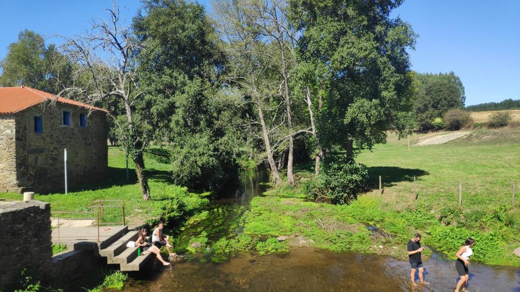 Vista de un área natural junto a un arroyo, donde un grupo de personas se relaja en el agua. Al fondo, se aprecia un edificio de piedra con ventanas azules y un techo rojo, rodeado de árboles y vegetación exuberante.