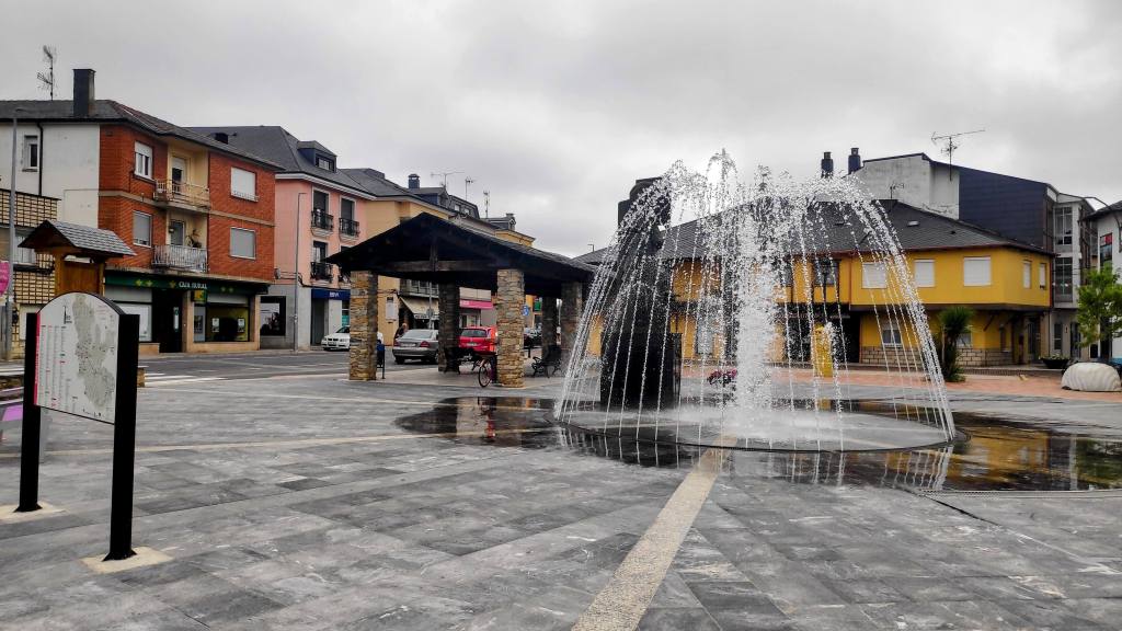 Etapa 25: Ponferrada-Villafranca del Bierzo - Caminando por Mallorca Plaza de Camponaraya, con una fuente central que emite chorros de agua, rodeada de edificios coloridos en un día nublado.