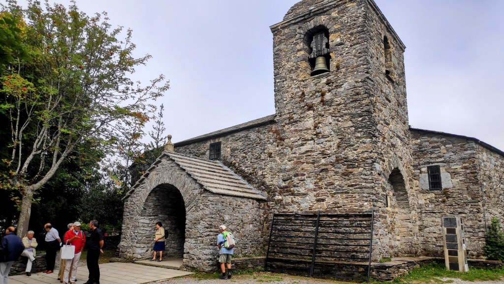 Grupo de personas reunidas frente a una iglesia de piedra con campanario en O Cebreiro, Galicia, rodeada de árboles y un ambiente natural.