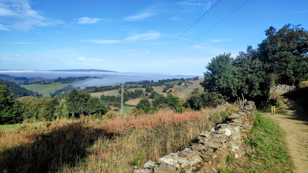 Vista panorámica de un valle en la ruta Triacastela-Sarria, con campos verdes y árboles, y un sendero que se extiende a lo largo del paisaje.