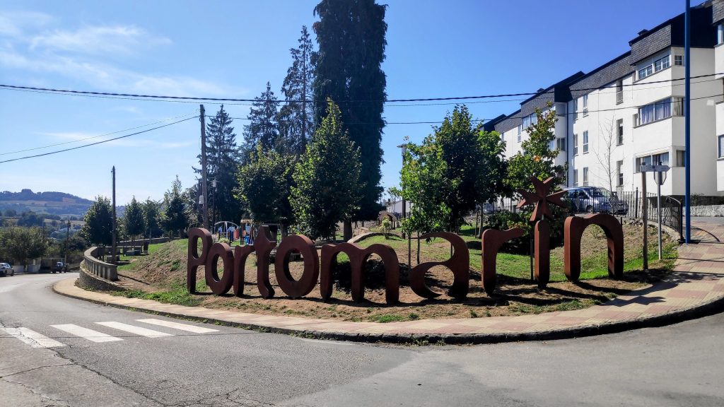 Cartel de bienvenida a Portomarín en un entorno urbano con árboles y edificios.