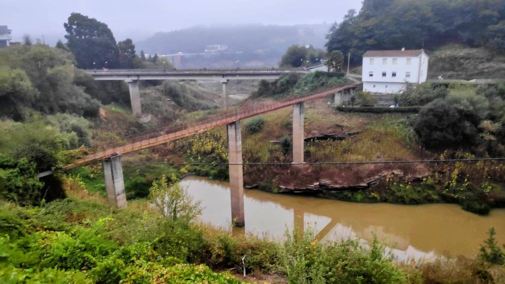 Vista de un puente de metal sobre un río en un entorno rural, con vegetación alrededor y un edificio blanco al fondo.