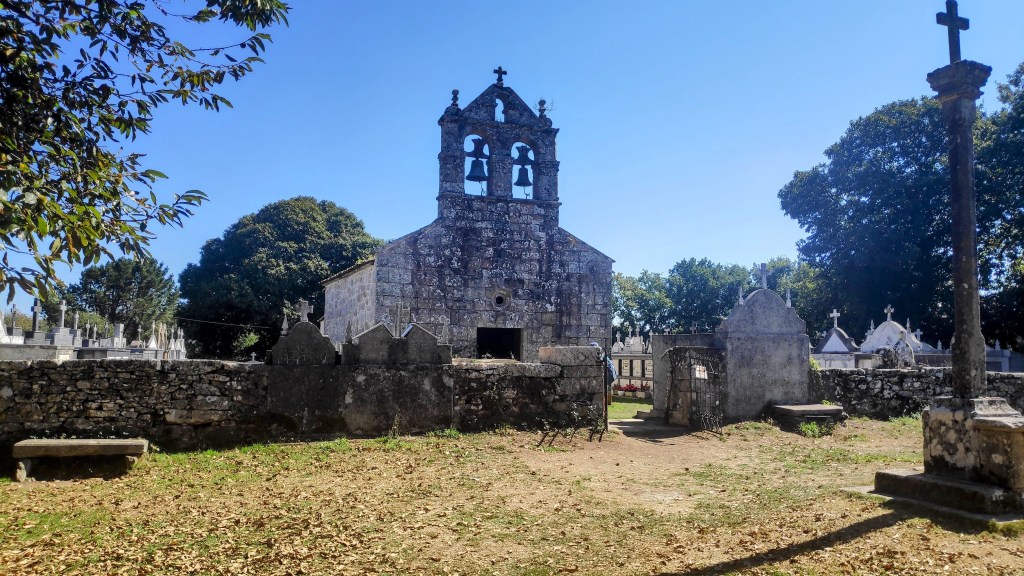 Iglesia con campanario y cementerio en un día soleado, rodeada de árboles y tumbas.