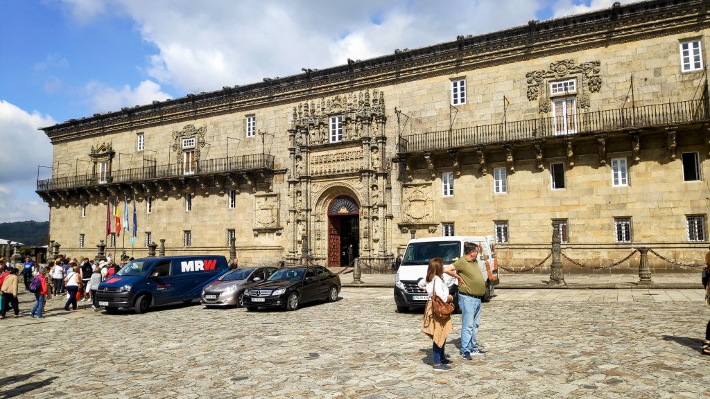 Vista del edificio del Hostal dos Reis Católicos en la Plaza del Obradoiro, Santiago de Compostela, con turistas y vehículos en primer plano.