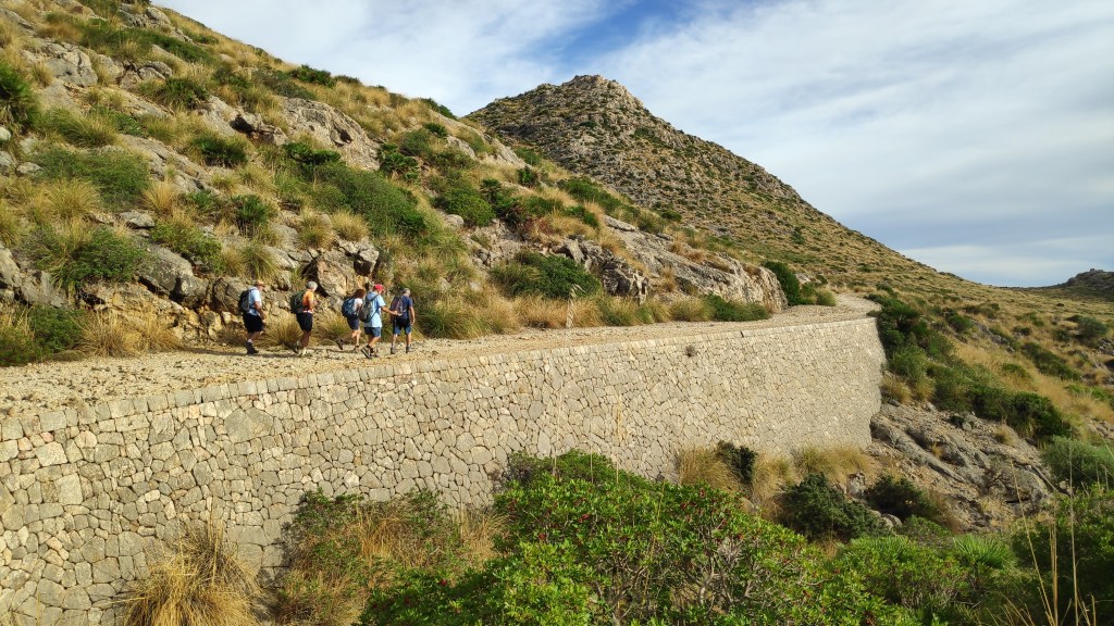 Grupo de senderistas caminando por un sendero de piedra junto a un terreno montañoso cubierto de vegetación en la ruta 'Puig de l'Águila'.