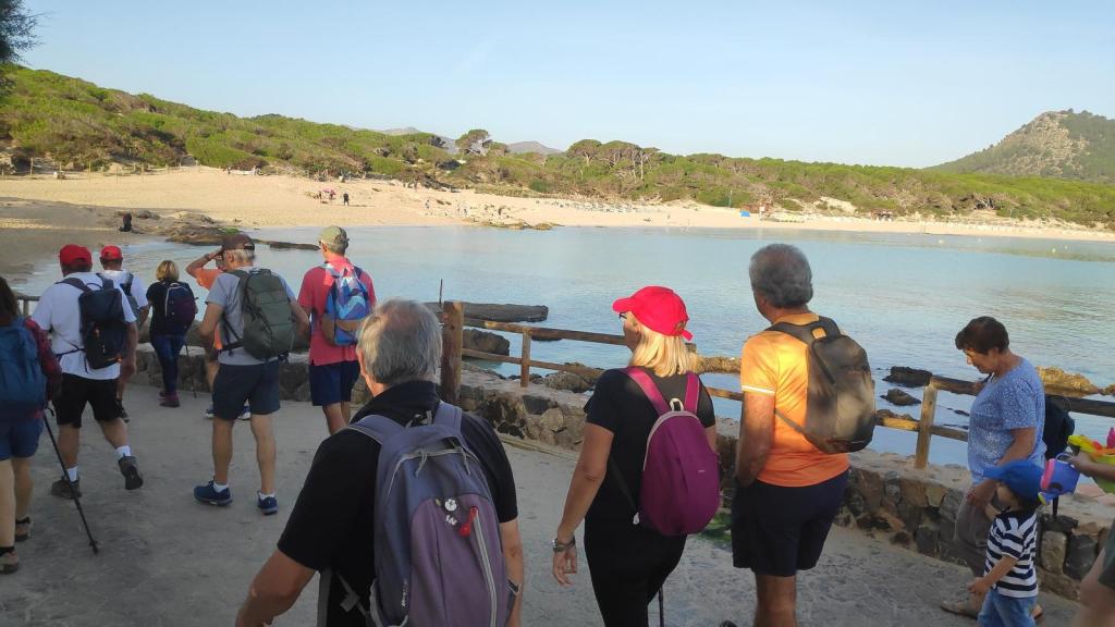Grupo de personas caminando hacia la playa de Cala Agulla, con árboles y montañas de fondo.