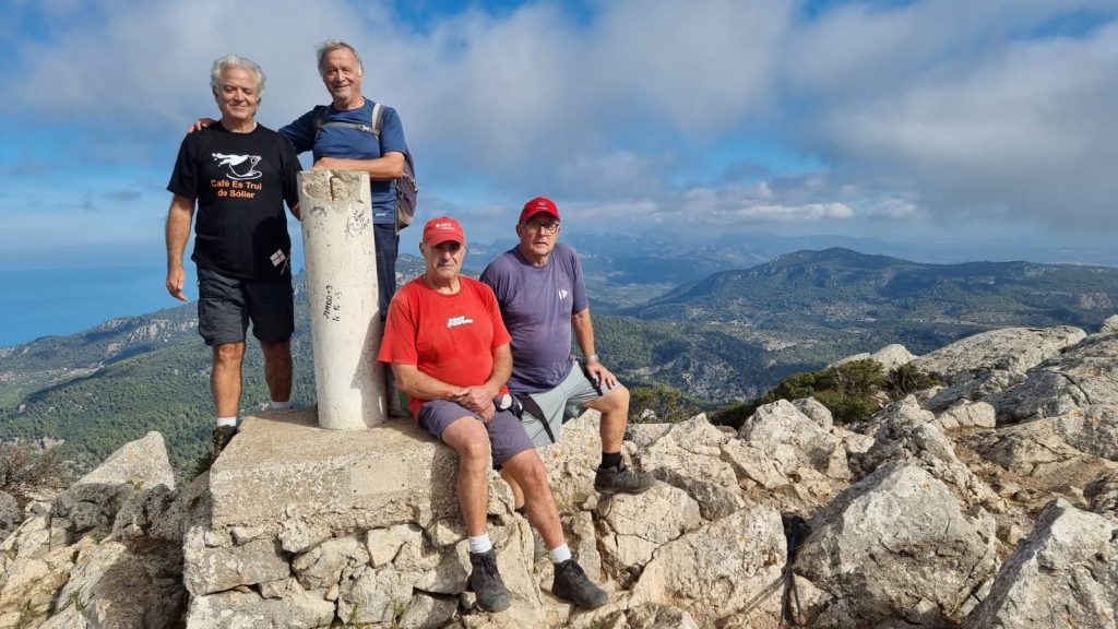 Puig de Galatzó por el Coll d’Estellencs - Caminando por Mallorca Cuatro personas posando en la cima del Puig de Galatzó con un vértice geodésico, rodeados de un paisaje montañoso y nublado.