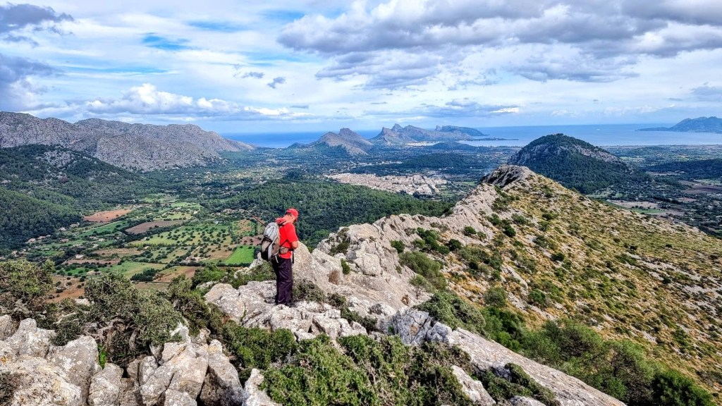 Vista panorámica desde la cima de la Serra de sa Coma, con un senderista de pie sobre rocas, observando el paisaje que incluye montañas y el mar en el fondo.