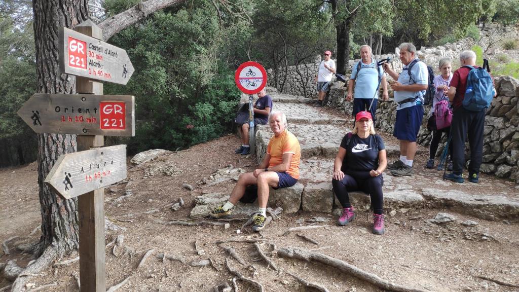 Castillo de Alaró desde Orient - Caminando por Mallorca Grupo de senderistas en un camino de montaña, con señales direccionales hacia el Castillo de Alaró y Orient, rodeados de vegetación y piedras.