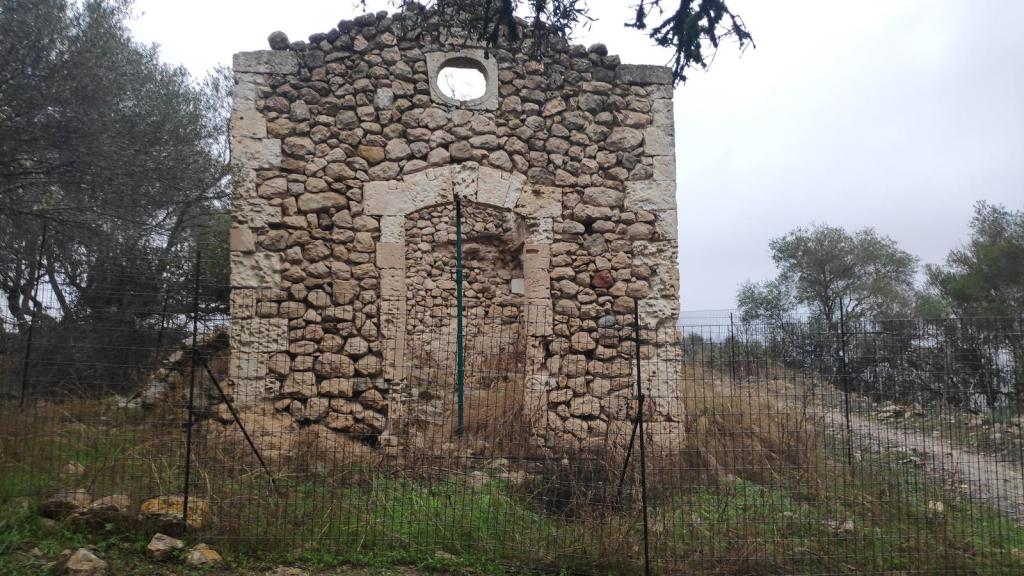 Ruinas de la Ermita de Sant Nofre, con paredes de piedra y una abertura en la parte superior, rodeadas de vegetación y rejas de metal.