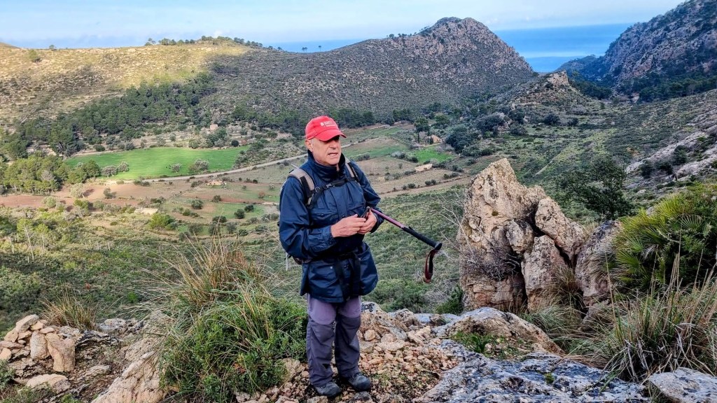 Hombre de pie en el Puig de ses Planes d’en Verd, con paisaje montañoso y mar al fondo, en la ruta ‘Puig de los Avencs-Coma Clova’.