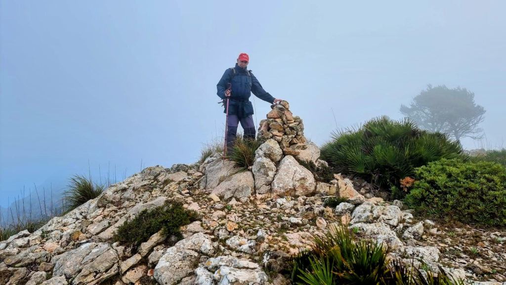 Persona en la cima del Puig des Avencs, rodeada de niebla, junto a una pequeña construcción de piedras en un terreno rocoso.