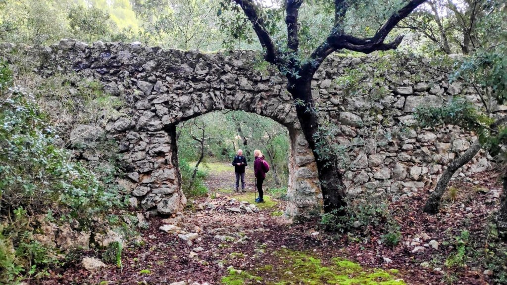 Arco de piedra antiguo en un sendero forestal, con dos caminantes al fondo rodeados de vegetación.