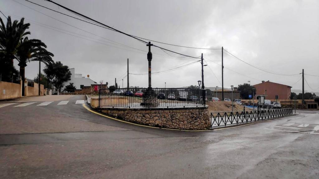 Vista de la Cruz d’en Raül con una zona de estacionamiento y edificios bajo un cielo nublado en Sant Joan, Mallorca.
