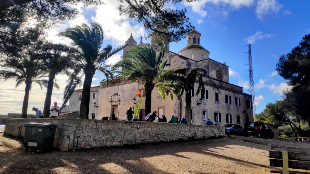 Vista del Santuario de Bonany rodeado de palmeras, con personas en el frente y coches aparcados, bajo un cielo parcialmente nublado.