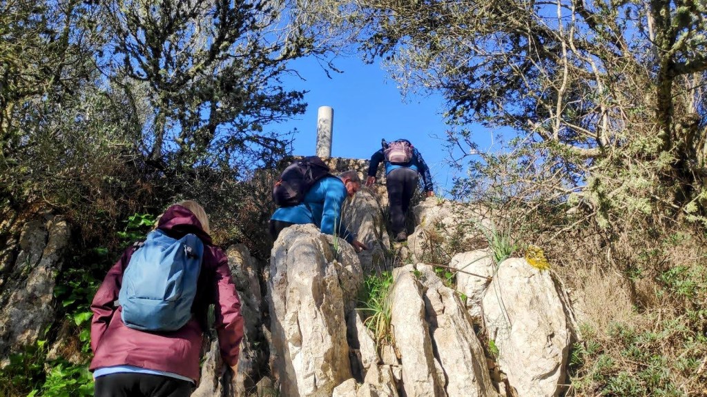 Tres personas ascendiendo por un terreno rocoso hacia la cima del Puig de Bonany, rodeadas de vegetación y bajo un cielo azul despejado.