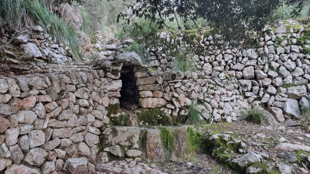 Fuente de mina restaurada hecha de piedra en seco, rodeada de paredes de piedra y vegetación en un sendero natural.