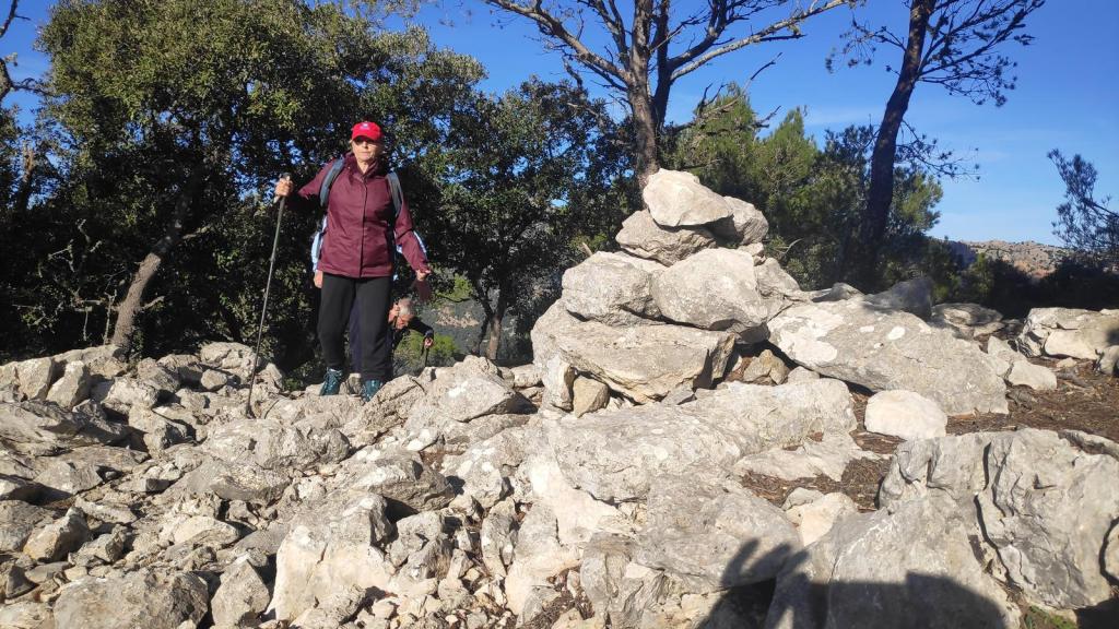 Senderista cruzando un terreno rocoso y accidentado, rodeado de árboles, mientras utiliza un bastón de trekking.