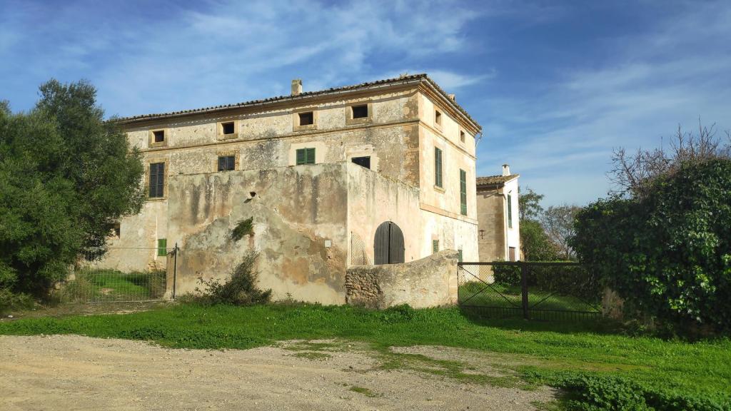 Vista de una antigua casa de campo con paredes desgastadas y un jardín verde que la rodea, bajo un cielo azul despejado.