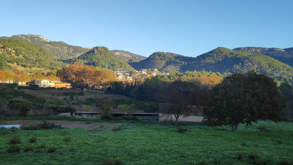 Panorama del pueblo de Puigpunyent con montañas al fondo y un cielo despejado. Se observan árboles de diversos colores y un campo verde en primer plano.