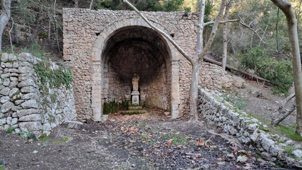 Mola de Son Pacs: Pasos - Caminando por Mallorca Fuente construida en una gruta natural, con una estatua en el centro, rodeada de paredes de piedra y vegetación.