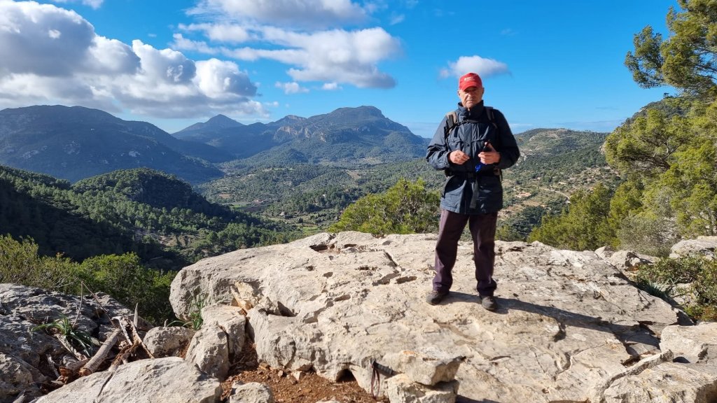 Hombre de pie en una roca, sosteniendo un objeto, con un paisaje montañoso y nublado de fondo.