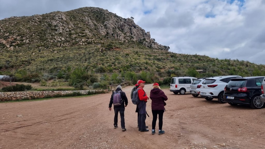 Grupo de personas preparándose para iniciar la ruta "Puig de los Avenc por el Coll de los Coloms" en un aparcamiento al aire libre, con montañas y vehículos estacionados en el fondo.