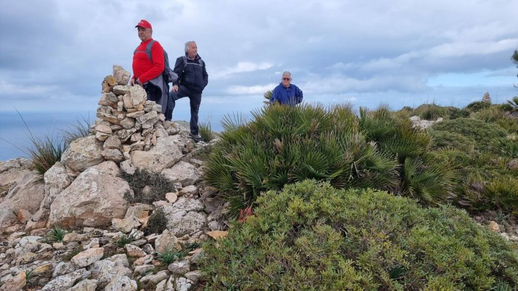 Grupo de tres personas en la cima del Puig des Avenc, rodeados de vegetación mediterránea y un paisaje montañoso.