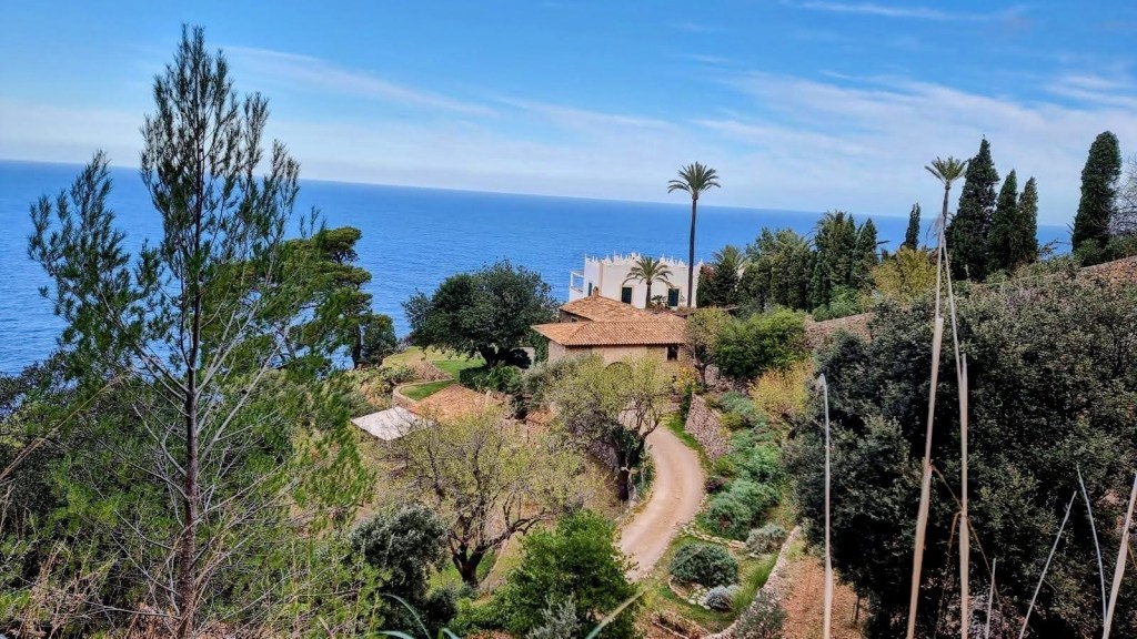Vista de una casa de s'Estaca rodeada de vegetación y palmeras, con el mar Mediterráneo al fondo.
