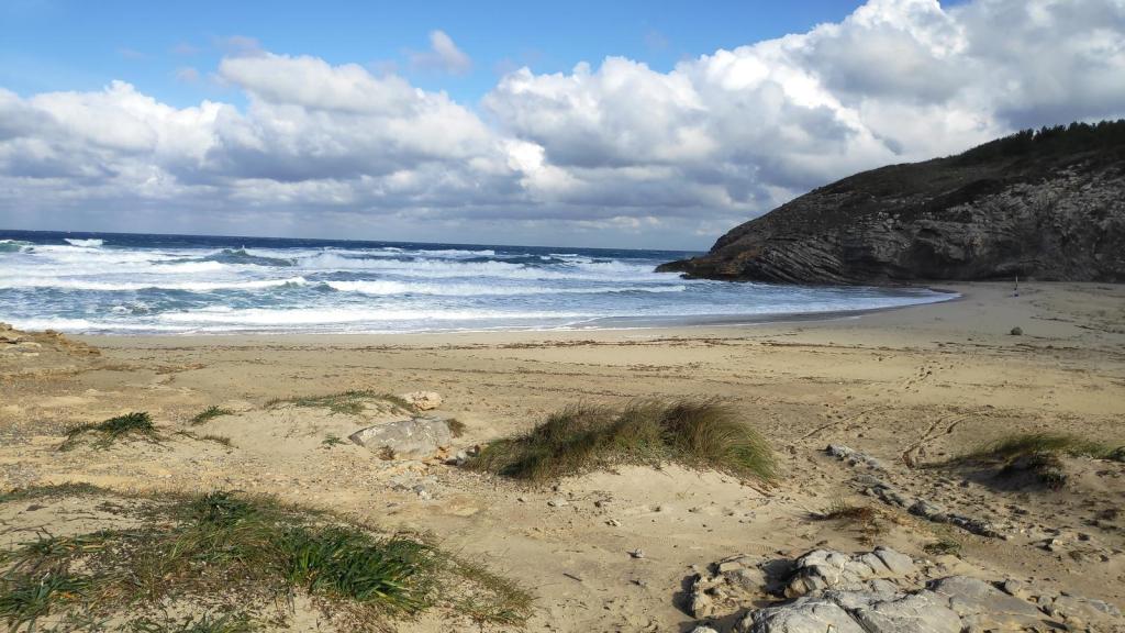 Vista de la playa con arena y olas del mar, rodeada de colinas y nubes en el cielo.
