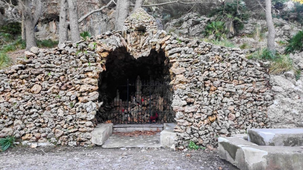 Cueva de piedra con una reja en la entrada, rodeada de vegetación y árboles, ubicada en un entorno natural.