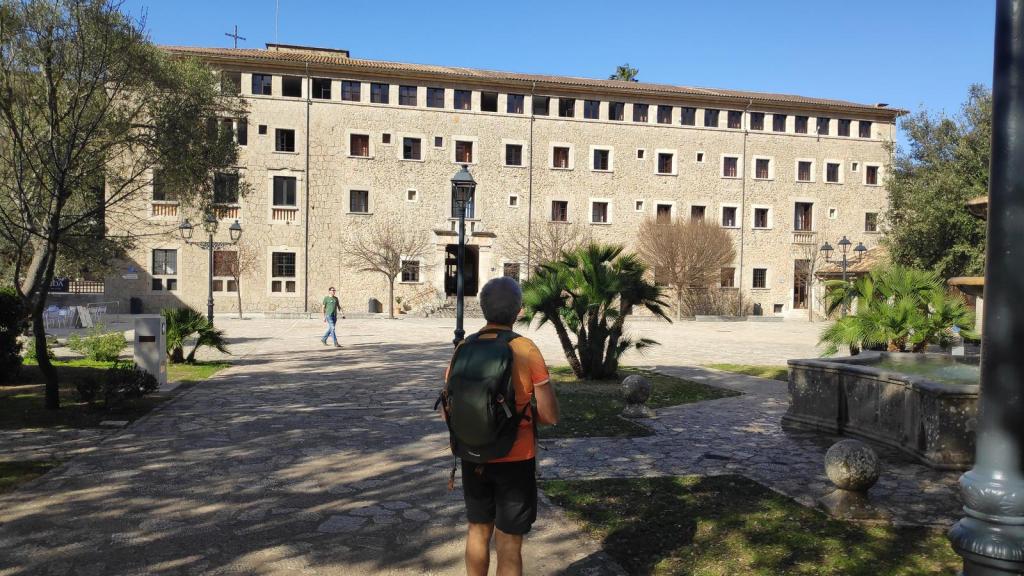 Vista del santuario de Lluc con un visitante en primer plano, rodeado de vegetación y con un cielo despejado.