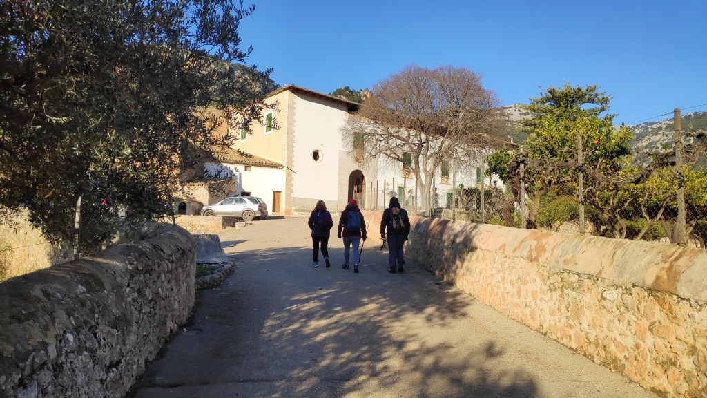 Tres excursionistas caminando por un camino de tierra, rodeados de árboles y casas típicas en el trayecto hacia Puig de sa Gubia, en Bunyola, Mallorca.