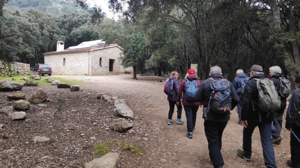 Grupo de senderistas caminando por un sendero en un bosque, con una casa de piedra visible al fondo.