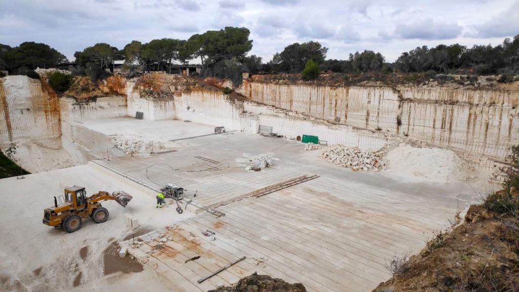 Vista panorámica de una cantera de mármol con maquinaria de construcción y bloques de piedra en el fondo, rodeada de vegetación y cielo nublado.