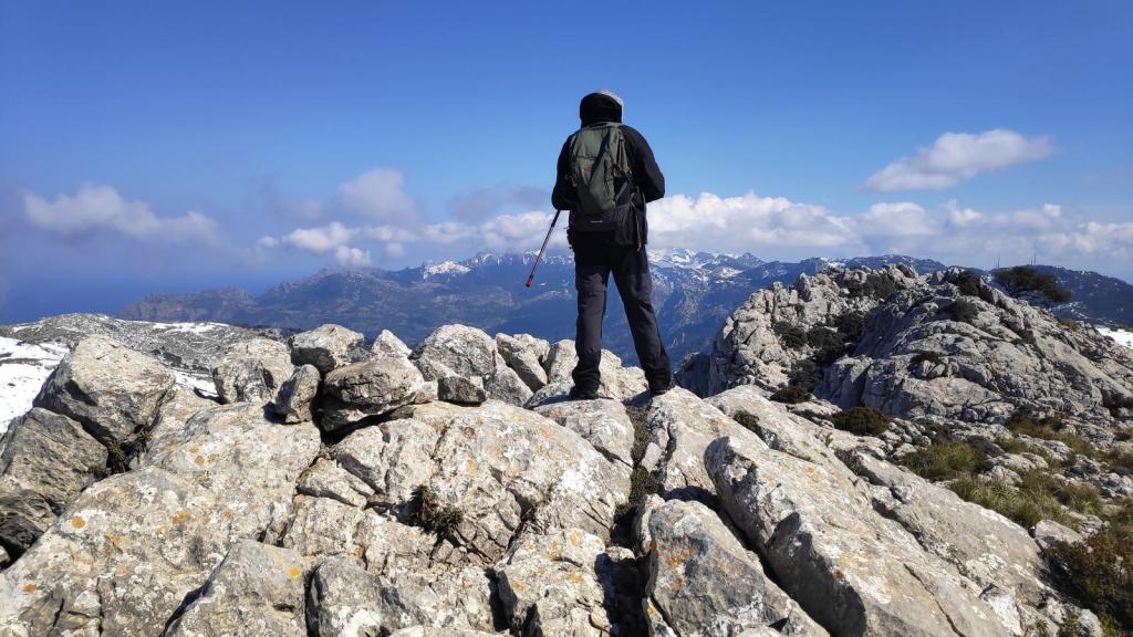 Un senderista de espaldas en la cima del Puig Teixot, rodeado de rocas y montañas nevadas bajo un cielo azul con algunas nubes.