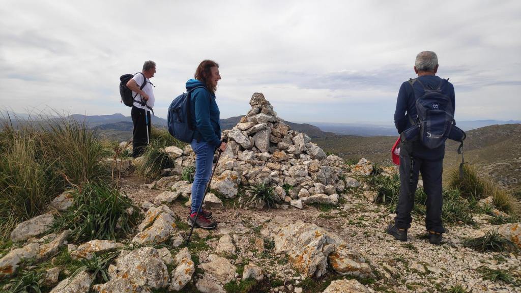 Puig del Porrassar desde s’Alquería Vella - Caminando por Mallorca Grupo de senderistas en la cima de Puig des Porrassar, con un hito de piedra a su lado y vistas panorámicas de la montaña.