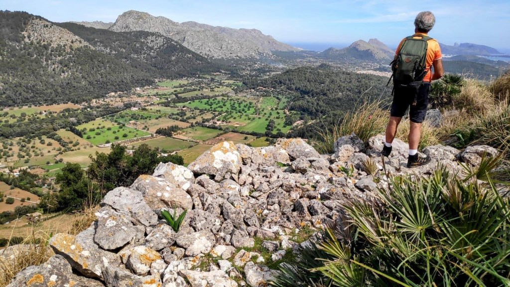 Serra de la Coma por la Regata de la Coma - Caminando por Mallorca Un excursionista de pie sobre una roca con vista panorámica de un valle verde y montañas de fondo en la ruta Serra de la Coma.